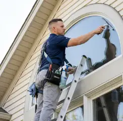 Professional window cleaner on a ladder using a squeegee to clean an exterior arched window, Chattanooga, TN.